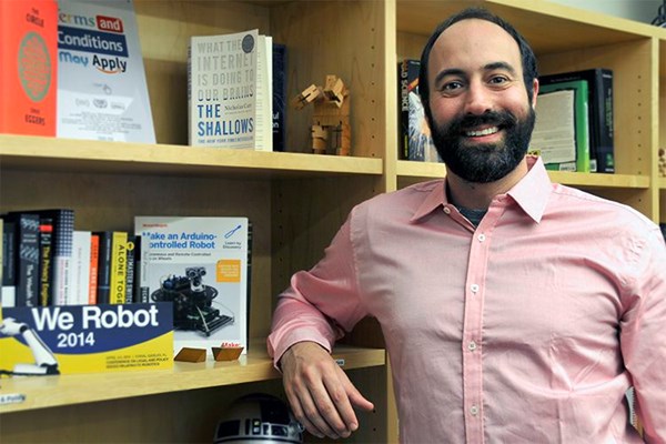 portrait of blog author, a man with dark hair and dark beard and pink shirt next to a bookcase