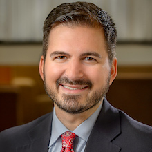Author Photo, a man with dark hair and beard wearing a suit, blue shirt and a red tie